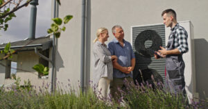 HVAC technician explains a large outdoor air conditioning unit to a couple outside a building.