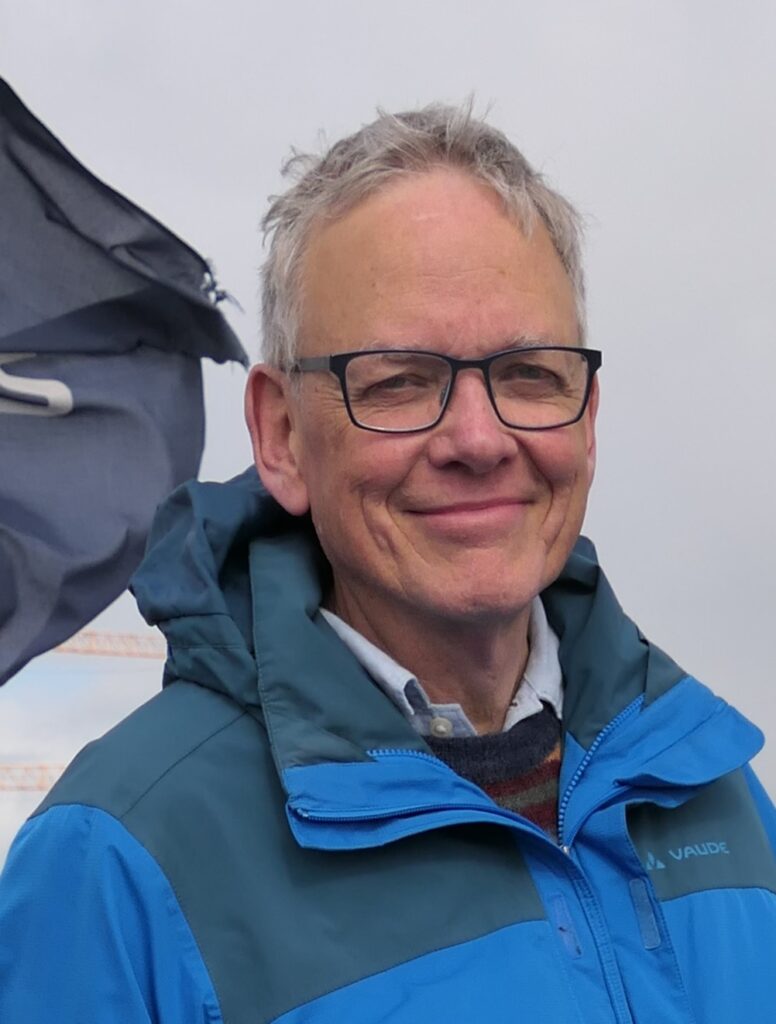 Smiling middle-aged man with gray hair and black glasses, wearing a blue and teal outdoor jacket outdoors against a cloudy sky.