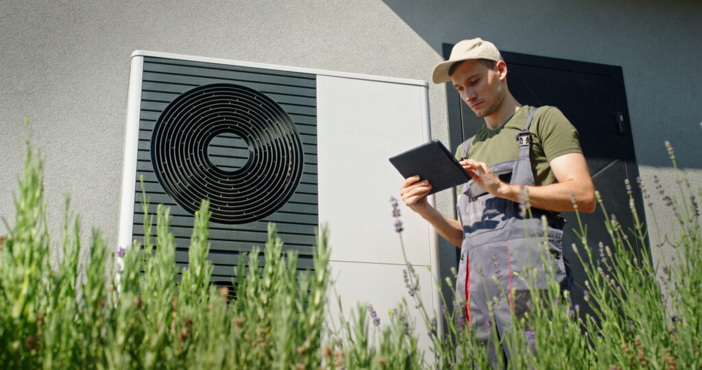 Maintenance worker in overalls and a beige cap uses a tablet beside an outdoor HVAC condenser unit.