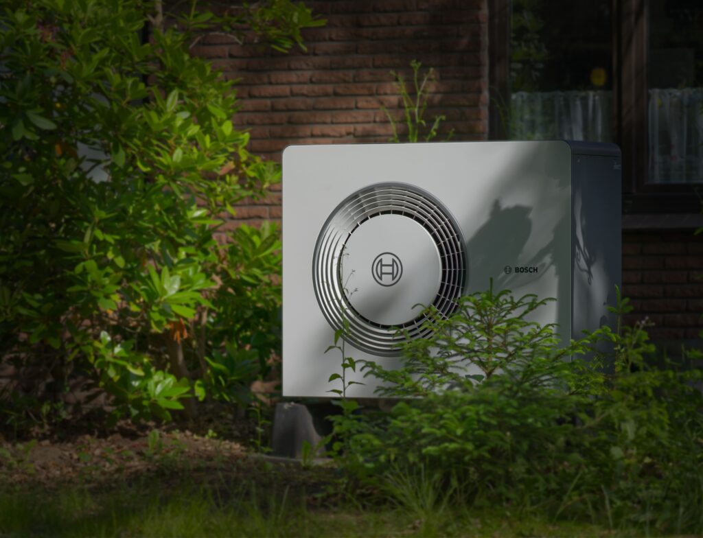 Outdoor Bosch air conditioner condenser unit placed beside a brick wall, with green bushes in the foreground.