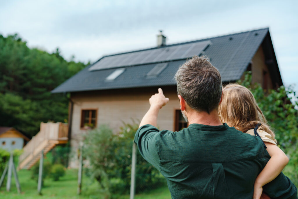 Back view of a man and a child pointing at a house with solar panels on the roof in a green yard